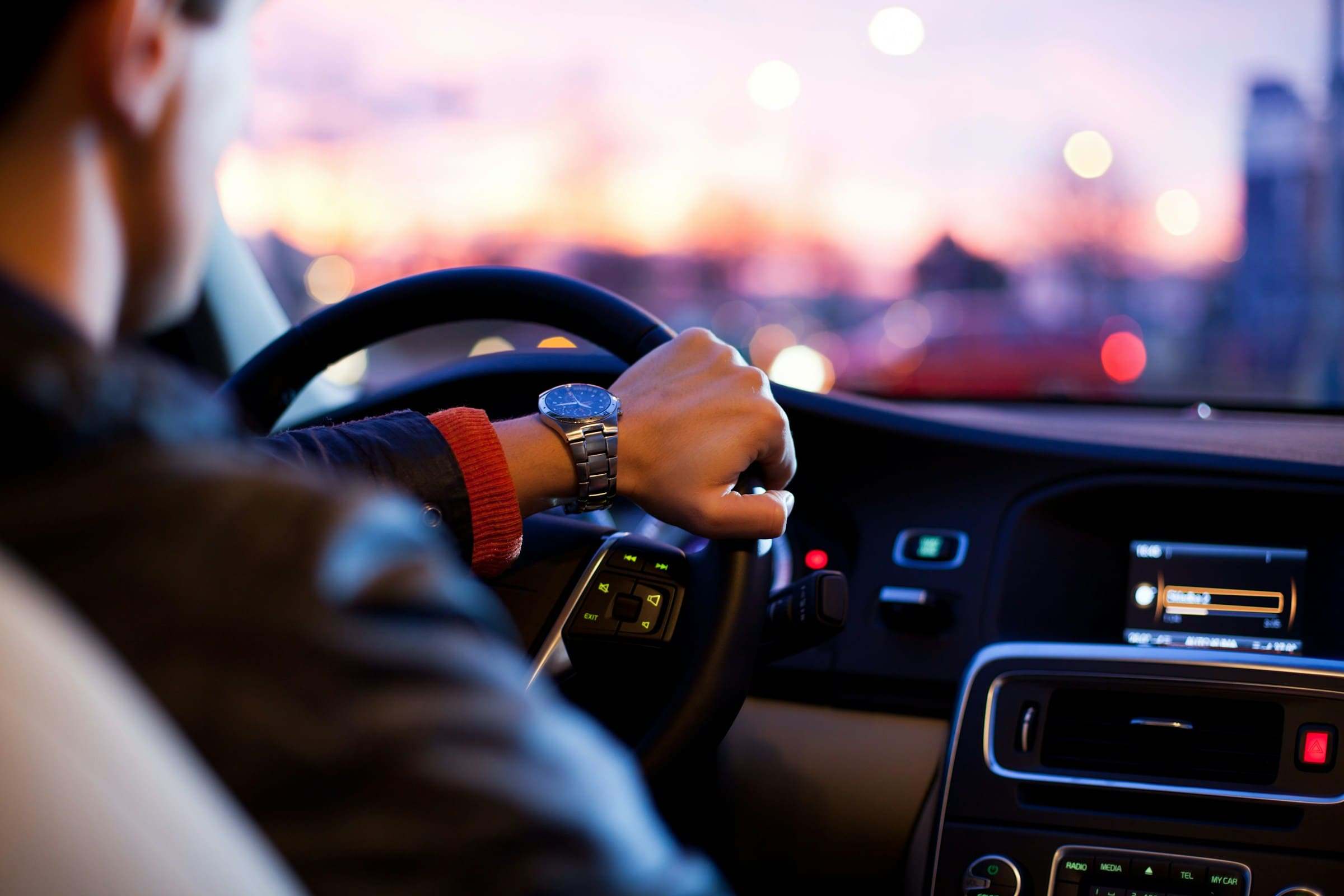 Ein Mann fährt ein Auto bei Sonnenuntergang. Man sieht seine Hand am Lenkrad, er trägt eine Uhr. Das Armaturenbrett und die Bedienelemente des Autos sind ebenfalls sichtbar.